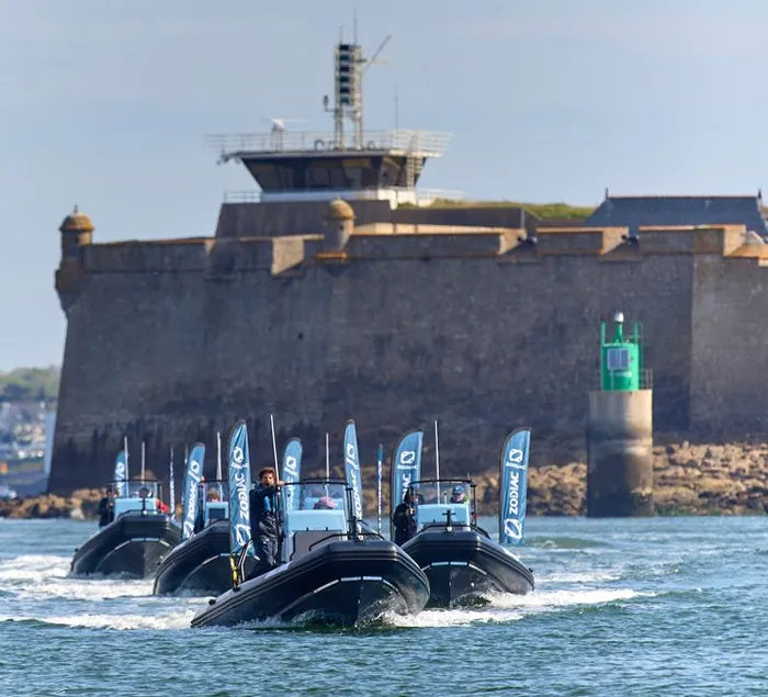 Formation of black Zodiac boats approaching a coastal fortress with flags raised